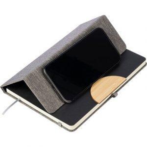 Carnet env. format A5 avec 80 pages lignées, ruban marque-page pour marquer facilement la dernière page ou les notes importantes, fermeture par élastique supplémentaire de couleur assortie, couverture pliable avec détail en bambou qui se transforme en support de téléphone, se glisse dans un sac à dos scolaire, produit fabriqué de RPET