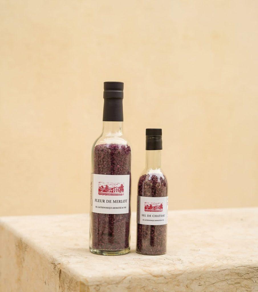 Deux flacons de sel violet alignés, main supportant les produits et fleurs violettes en arrière‑plan