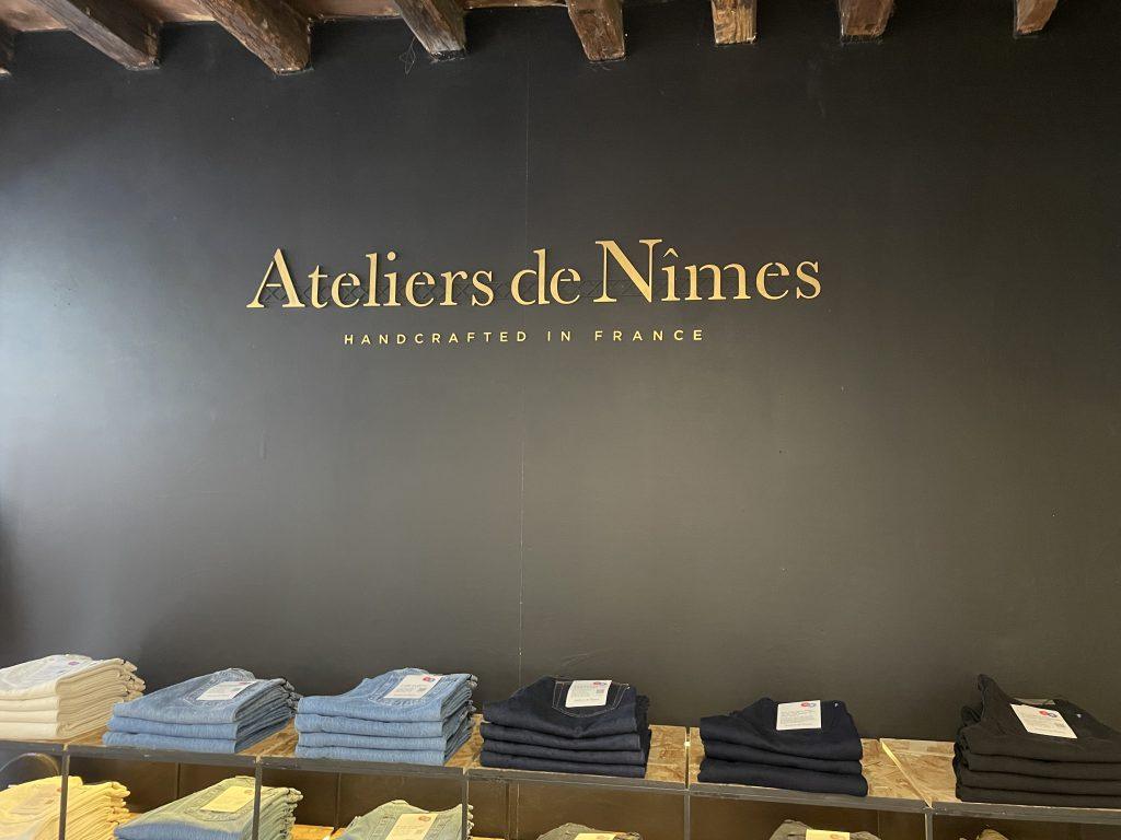 Mur noir décoratif avec lettrage doré 'Ateliers de Nîmes' au-dessus d’une table présentant des jeans