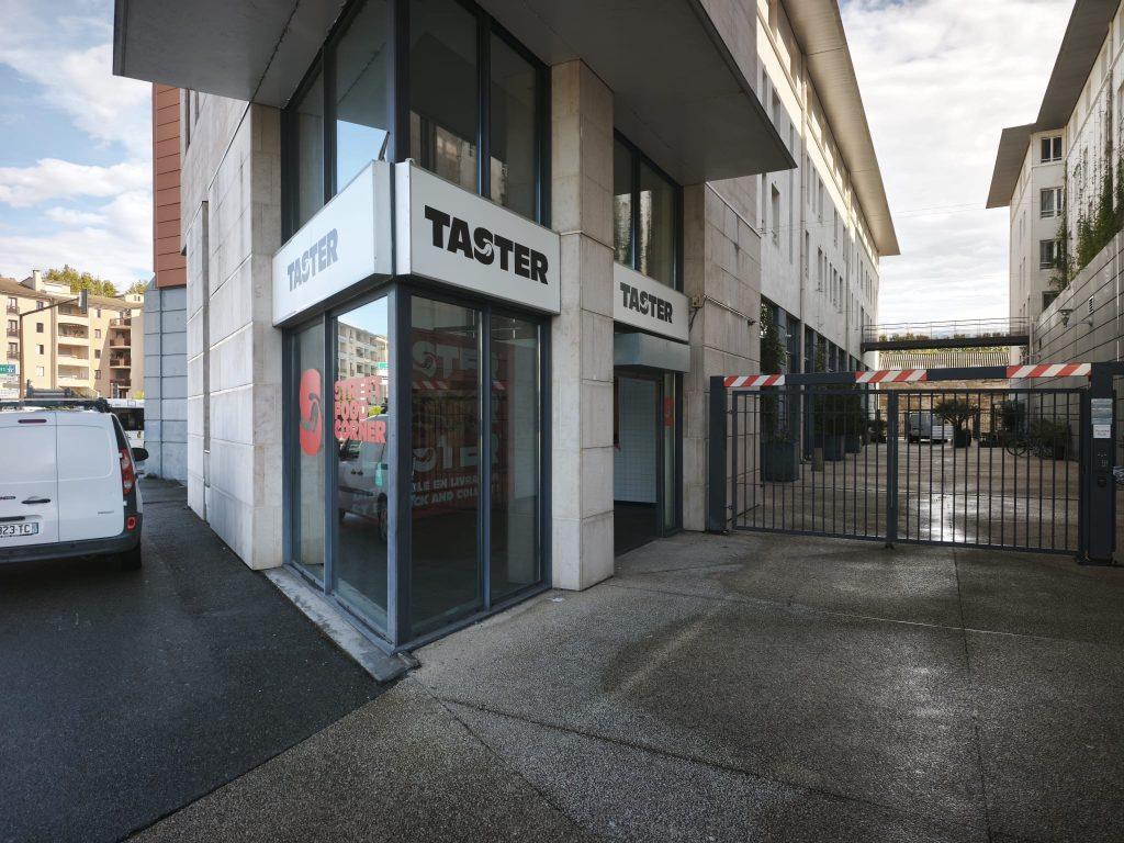 Entrée en angle du magasin Taster avec enseigne blanche et baie vitrée